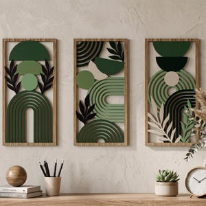Op de afbeelding: Drie ingelijste wandkunstwerken met abstracte geometrische ontwerpen in tinten groen, zwart en beige. Elk stuk heeft gelaagde vormen, waaronder bogen, cirkels en bladvormige uitsparingen, geplaatst in een natuurlijke houten lijst. De kunstwerken worden tentoongesteld op een neutraal getinte muur.