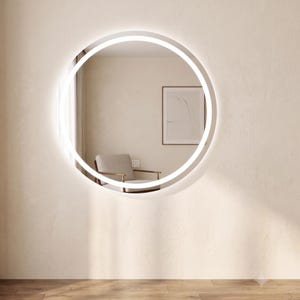 Peut inclure: Un miroir rond éclairé par une lumière LED blanche. Le miroir est fixé sur un mur beige, reflétant une pièce avec une chaise et une œuvre d'art. Le sol est en bois.