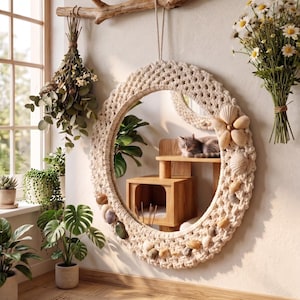 Peut inclure: Un miroir en macramé avec un lit pour chat et des accents de coquillages. Le miroir ovale est entouré de macramé crème et orné de coquillages et d'une fleur en bois. Un chat gris se repose sur une étagère en bois à l'intérieur du miroir.