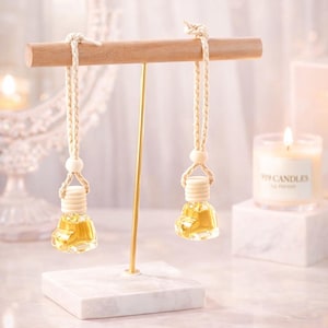 May include: Two small, clear glass bottles filled with yellow liquid hang from a wooden bar. The bottles are suspended by beige rope and wooden beads. The bar is supported by a gold metal stand on a white marble base. The text "919 CANDLES" is at the bottom.