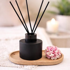 May include: A black essential oil diffuser with black reeds, sitting on a wooden tray. A pink flower is next to the diffuser. The background is blurred, with a lit candle and greenery.