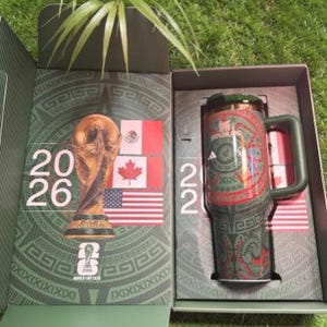 May include: Green and red insulated travel mug with a handle, featuring a decorative pattern and the Adidas logo. The mug is presented in a box with the FIFA World Cup trophy and flags of Mexico, Canada, and the United States.
