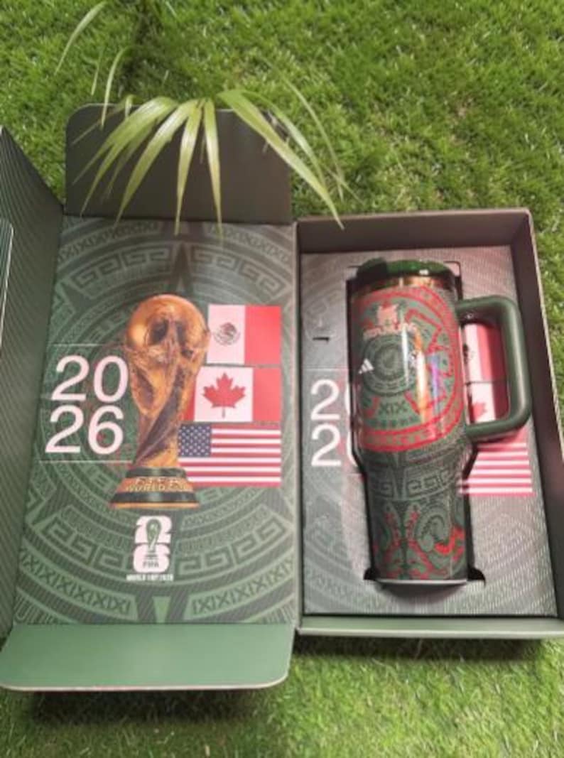 May include: Green and red insulated travel mug with a handle, featuring a decorative pattern and the Adidas logo. The mug is presented in a box with the FIFA World Cup trophy and flags of Mexico, Canada, and the United States.