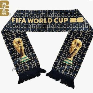 May include: A black scarf with gold accents and fringe. The scarf features the text "FIFA WORLD CUP 26" in gold, along with a repeating pattern and images of the World Cup trophy. The FIFA logo is visible.