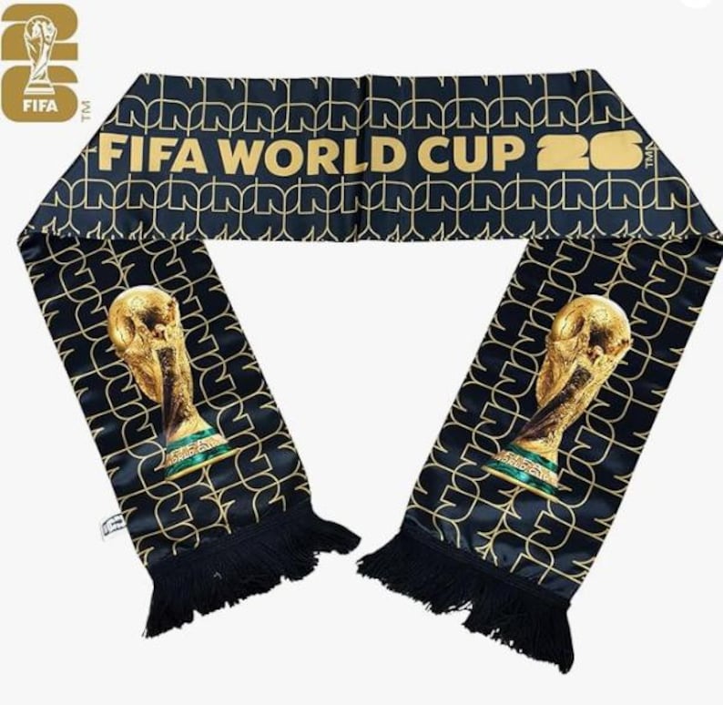 May include: A black scarf with gold accents and fringe. The scarf features the text "FIFA WORLD CUP 26" in gold, along with a repeating pattern and images of the World Cup trophy. The FIFA logo is visible.