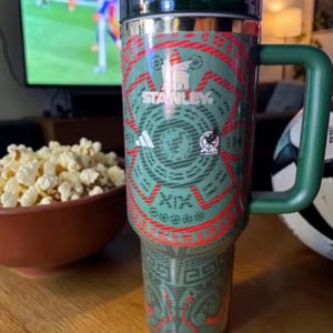 May include: A green and red Stanley travel mug with a handle, featuring a design with the Adidas logo and the Mexican national team emblem. The mug is on a wooden table, with a bowl of popcorn and a soccer ball nearby.