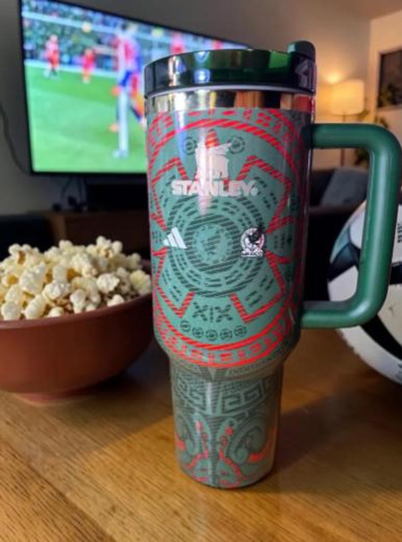 May include: A green and red Stanley travel mug with a handle, featuring a design with the Adidas logo and the Mexican national team emblem. The mug is on a wooden table, with a bowl of popcorn and a soccer ball nearby.