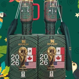 May include: Two Stanley travel mugs with red handles and lids, featuring an Aztec-inspired design and the Adidas logo. They sit atop boxes displaying the FIFA World Cup trophy, flags of the USA, Canada, and Mexico, and the year 2026.