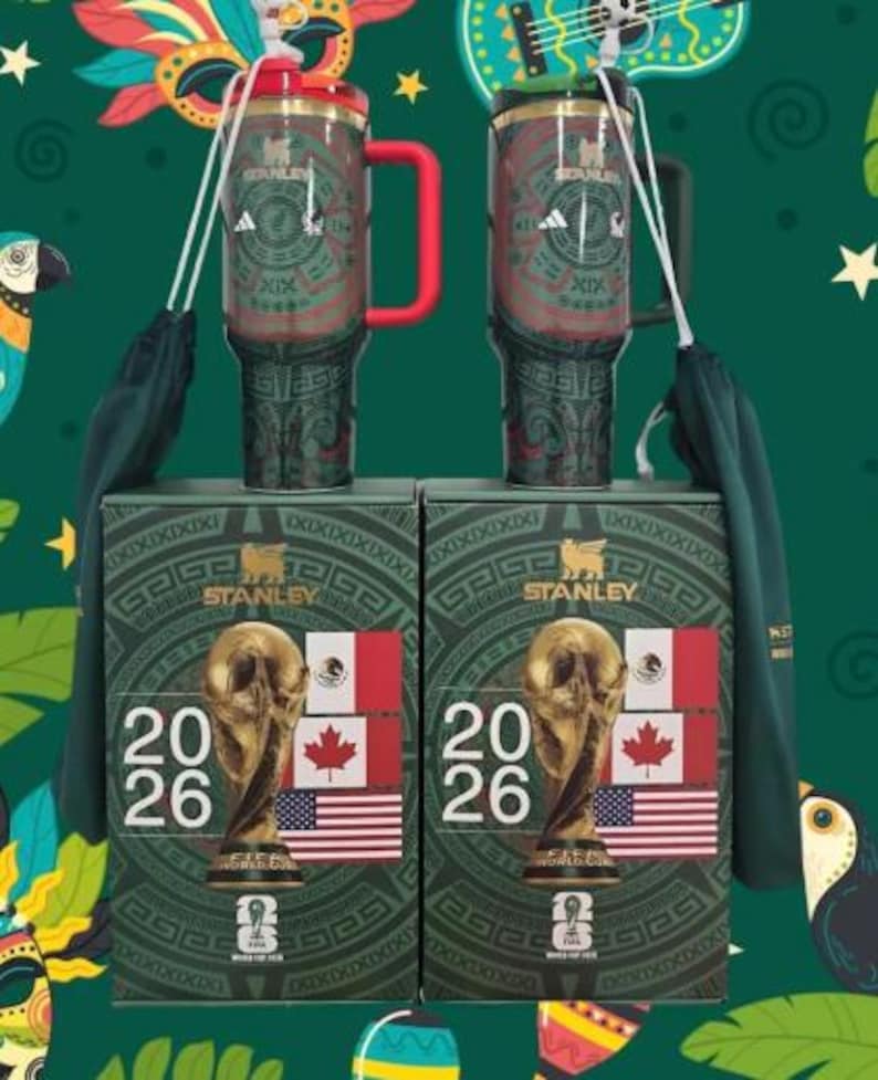May include: Two Stanley travel mugs with red handles and lids, featuring an Aztec-inspired design and the Adidas logo. They sit atop boxes displaying the FIFA World Cup trophy, flags of the USA, Canada, and Mexico, and the year 2026.