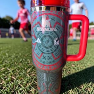 May include: A stainless steel Stanley travel mug with a red handle and lid. The mug has a teal and red Aztec-inspired design with the Adidas and Stanley logos. The background shows a soccer field with children playing.