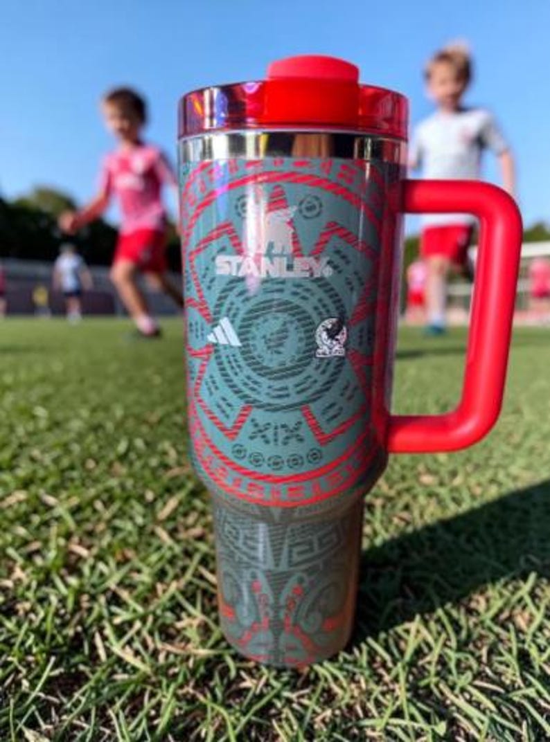 May include: A stainless steel Stanley travel mug with a red handle and lid. The mug has a teal and red Aztec-inspired design with the Adidas and Stanley logos. The background shows a soccer field with children playing.