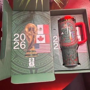 May include: A green and red travel mug with a red handle and lid, inside a box with a green and gold design. The box features the text "2026" and flags of Mexico, Canada, and the United States. The mug has a repeating Aztec-inspired pattern.
