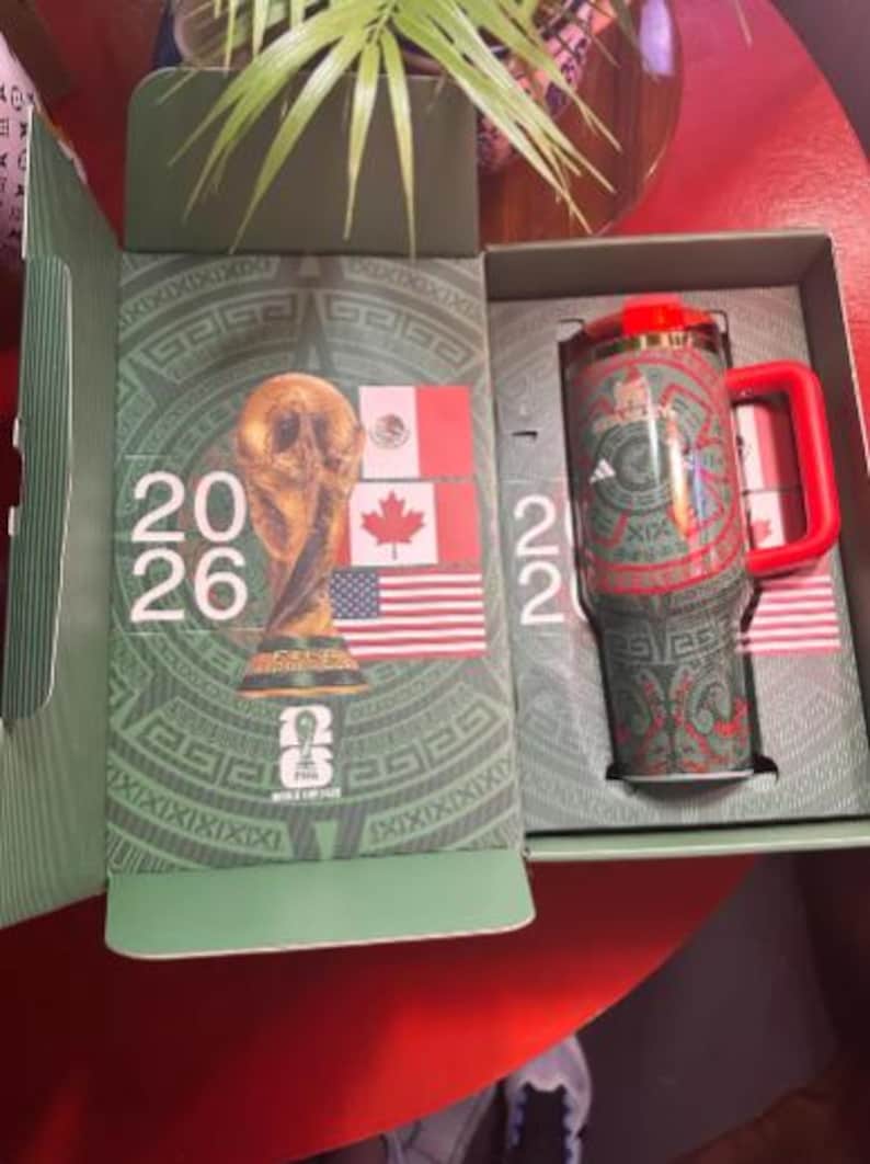 May include: A green and red travel mug with a red handle and lid, inside a box with a green and gold design. The box features the text "2026" and flags of Mexico, Canada, and the United States. The mug has a repeating Aztec-inspired pattern.