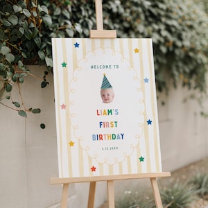 Op de afbeelding: Een welkomstbord voor een verjaardag op een houten ezel. Het bord heeft een gestreepte achtergrond, kleurrijke sterren en de tekst "Welcome to Liam's First Birthday". Een foto van een baby met een feesthoedje is afgebeeld.