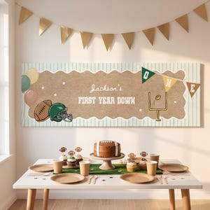 Puede incluir: Una decoración de fiesta con temática de fútbol que incluye un cartel con el texto Jackson's FIRST YEAR DOWN, un pastel marrón con forma de balón, cupcakes, platos de papel y vasos de color tostado sobre una mesa con un camino verde.