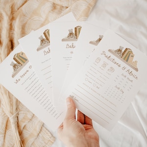 May include: A set of white paper baby shower game cards featuring a brown teddy bear sitting on a shelf with books. The cards include printed text such as Who Knows Mommy, Baby Predictions and Advice, and lines for writing responses in brown ink.