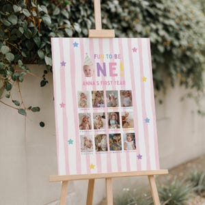 以下が含まれることがあります： 木製のイーゼルには、「FUN TO BE ONE！」と「ANNA'S FIRST YEAR」という文字が入ったピンクと白のストライプのポスターが飾られています。ポスターには赤ちゃんの写真のコラージュとカラフルな星のアクセントが施されています。