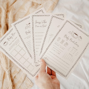 Puede incluir: Un conjunto de tarjetas de juego para baby shower de color crema con texto marrón oscuro y bordes festoneados. Las tarjetas incluyen títulos como Baby Bingo, Guess Who Mom or Dad y Baby Predictions, con ilustraciones de tazas.