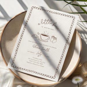 May include: A baby shower invitation with a beige background and brown text. The text reads "a little bean is brewing!" with illustrations of a coffee cup, croissant, and baby socks. The invitation is on a beige plate.