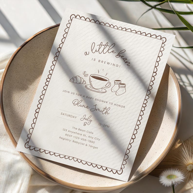 May include: A baby shower invitation with a beige background and brown text. The text reads "a little bean is brewing!" with illustrations of a coffee cup, croissant, and baby socks. The invitation is on a beige plate.