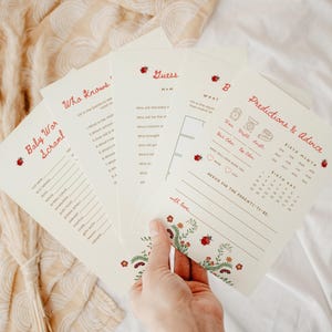 May include: A set of cream-colored baby shower game cards featuring red text and ladybug illustrations. The cards include titles like Baby Word Scramble, Who Knows Mommy Best, and Predictions & Advice, with lines for writing answers and dates.
