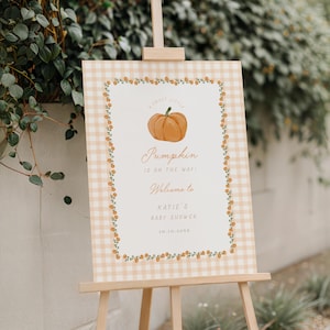 Peut inclure: Une pancarte rectangulaire sur un chevalet en bois avec une bordure à carreaux orange clair et blanc. Le centre présente une illustration de citrouille avec le texte : A SWEET LITTLE Pumpkin IS ON THE WAY! Welcome to KATIE'S BABY SHOWER 10.10.20XX.