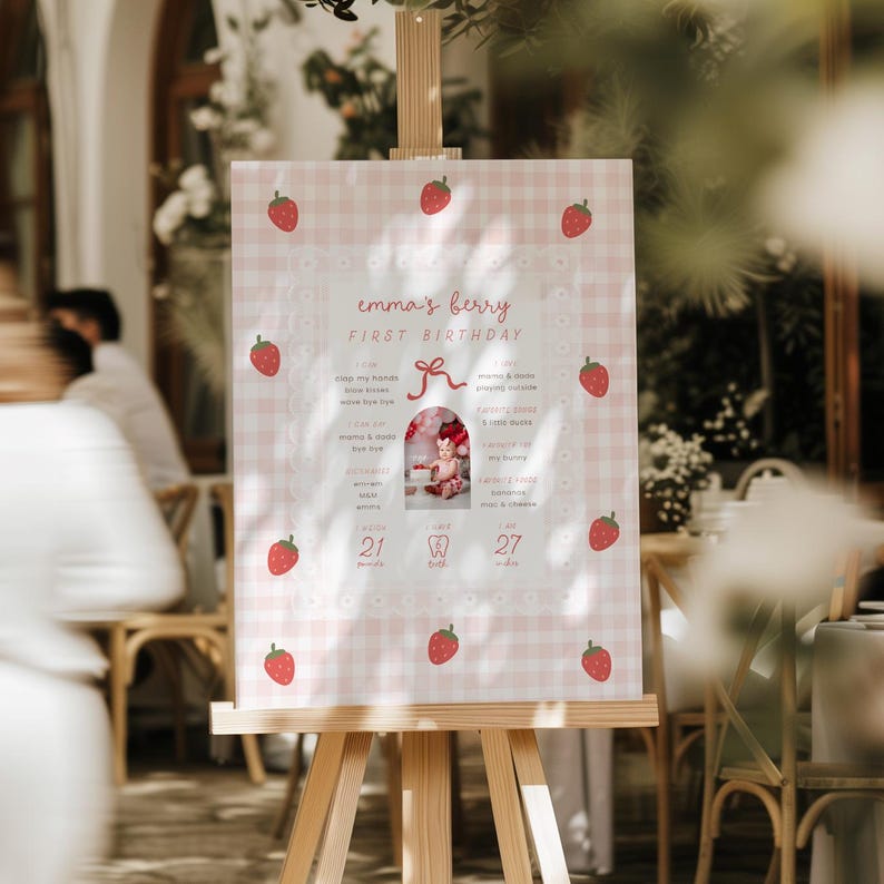 May include: A pink and white gingham-patterned sign on a wooden easel. The sign reads "emma's berry FIRST BIRTHDAY" with strawberry illustrations. A photo of a baby is in the center. The sign includes facts about the baby.