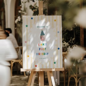 Può includere: Un cavalletto in legno espone un cartello con la foto di un bambino e la scritta "Benvenuti al Primo Compleanno di Liam". Il cartello ha uno sfondo a strisce con stelle colorate e un bordo decorativo. È indicata anche la data 3.15.20XX.