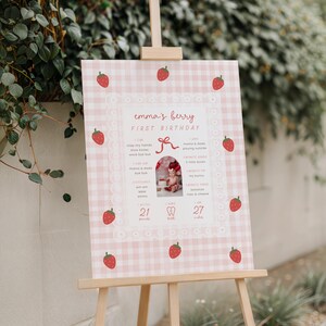 May include: A pink and white gingham-patterned sign on a wooden easel, featuring the text "emma's berry FIRST BIRTHDAY." The sign includes a photo and strawberry illustrations. It lists milestones and the baby's weight as 21 pounds.