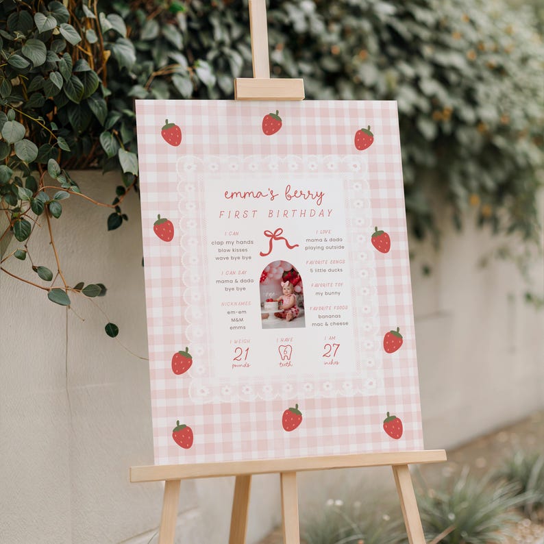 May include: A pink and white gingham-patterned sign on a wooden easel, featuring the text "emma's berry FIRST BIRTHDAY." The sign includes a photo and strawberry illustrations. It lists milestones and the baby's weight as 21 pounds.