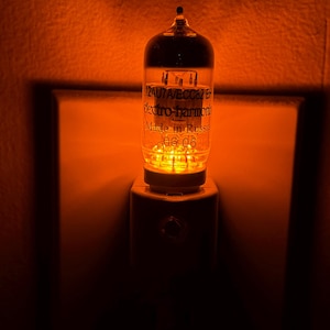 May include: A vacuum tube night light emitting a warm orange glow. The glass tube is labeled 12AU7A/ECC82 EH, electro-harmonix, and Made in Russia 06 06. The cylindrical tube is mounted on a white plastic base plugged into a wall outlet.