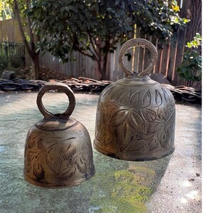 May include: Two brass bells of different sizes, each featuring a circular handle at the top and an engraved leaf pattern covering the exterior surface. The metal has a weathered, muted gold finish with dark etched details highlighting the foliage.