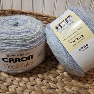 May include: Two skeins of Caron Cloud Cakes yarn in shades of blue, green, and white. Each skein is labeled with the brand name and product details. One skein is partially unraveled, revealing the yarn's texture. The label indicates each skein weighs 8 oz.