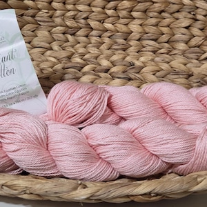 May include: A woven basket holds pink yarn skeins and a label that reads "Radiant Cotton." The yarn is a soft, light pink color. The basket is made of woven natural fibers.
