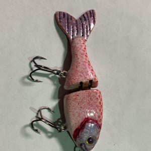 May include: A two-piece fishing lure shaped like a fish, primarily white with pink speckles. The tail is purple and the lure has two treble hooks. The eye is silver with a yellow center.