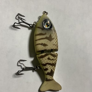 May include: A two-piece fishing lure in the shape of a fish. The lure is tan with brown markings and a reflective eye. Two treble hooks are attached. The lure is designed to attract fish.