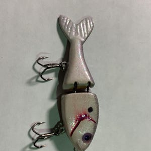 May include: A two-piece fishing lure in a fish shape, with a pearlescent finish. The lure has a tail, a head with a purple eye, and two treble hooks. The lure is designed to attract fish.
