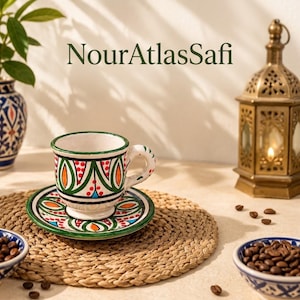 May include: A ceramic coffee cup and saucer set featuring hand-painted green, orange, and blue geometric patterns on a white background. The text NourAtlasSafi is visible above the cup. The set is accompanied by two small bowls of coffee beans.