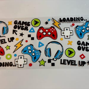 May include: A clear sticker sheet featuring colourful gaming-themed graphics including red and blue game controllers, blue headphones, yellow lightning bolts, and green play buttons. The text reads "GAME OVER", "LEVEL UP", and "LOADING.." in black.