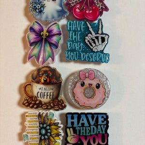 May include: A collection of eight decorative acrylic badges featuring various designs including a ghost with a blue bow, cherries with a leopard print bow, a purple bow, a skeleton hand with the text HAVE THE DAY YOU DESERVE, a dog in a coffee mug, a pink donut, a sunflower with text, and a sign with the text HAVE THE DAY YOU DESERVE.
