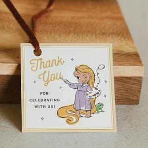 Puede incluir: Una etiqueta de regalo cuadrada blanca con un cordón de terciopelo marrón que presenta el texto dorado Thank You FOR CELEBRATING WITH US! y una ilustración de un personaje de pelo largo con vestido morado sosteniendo un pincel y una paleta junto a un pequeño lagarto verde.
