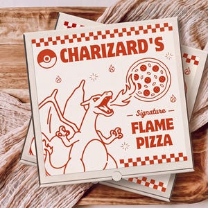 May include: A square white cardboard pizza box featuring red graphics. The design includes a red checkered border, a Poké Ball icon, and a line drawing of a dragon-like creature breathing fire toward a pizza. Text reads Charizard's Signature Flame Pizza.