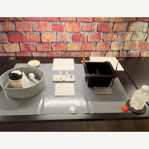 Puede incluir: Una configuración de manualidades sobre una superficie gris, con un fondo de pared de ladrillo. Incluye un recipiente gris con suministros, paletas de pintura, una bandeja de pintura negra, una botella de agua y un posavasos rojo. La configuración sugiere una actividad de pintura o manualidades.