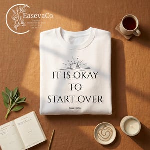 Puede incluir: Una sudadera blanca de cuello redondo con un gráfico de líneas negras de un sol naciente y el texto "IT IS OKAY TO START OVER" impreso en una fuente serif. El nombre de la marca "EasevaCo" aparece en una fuente más pequeña debajo.