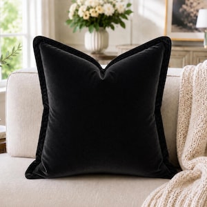 Könnte beinhalten: Ein großes, quadratisches schwarzes Samtkissen mit kontrastierendem dunklen Besatz. Das Kissen liegt auf einem cremefarbenen Sofa, mit einer gestrickten Decke über dem Arm. Der Hintergrund umfasst eine Vase mit Blumen und ein gerahmtes Bild.