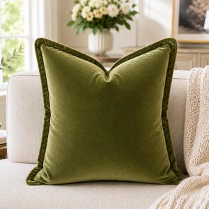 May include: An olive green velvet cushion with a darker green trim, placed on a cream sofa. A knitted throw is draped over the sofa arm. The cushion is a decorative item for the home, adding a touch of colour.