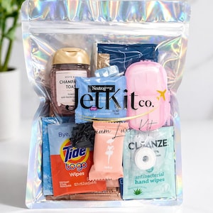 May include: A clear, holographic zip-lock bag filled with travel-sized toiletries and personal care items. Includes a small bottle of Champagne Toast, Neutrogena wipes, Tide to Go wipes, and antibacterial hand wipes. The bag is labeled "JetKit co." and "Premium Luxe Kit."