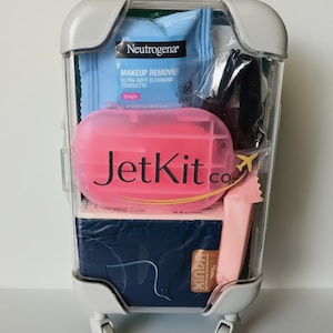 May include: A clear, miniature suitcase filled with travel-sized toiletries. The case has a white frame and wheels. Inside are a blue Neutrogena makeup remover towelette, a pink JetKit co. case, and other personal care items.