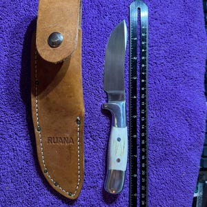 May include: A hunting knife with a stainless steel blade and a white handle with brown spots. The knife is next to a brown leather sheath with the word "RUANA" embossed on it. A ruler is next to the knife.