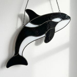 May include: A stained glass orca suncatcher featuring black and white glass segments joined by dark metal solder lines. The orca is depicted in a swimming pose with a dorsal fin, pectoral fin, and tail fluke, suspended by a thin metal chain.
